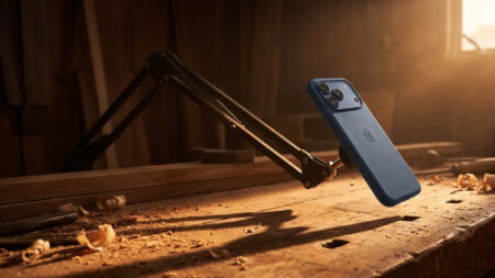 iPhone 18 Pro: código do iOS vaza recursos de câmera Smartphone em suporte articulado de metal em uma bancada de marcenaria rústica, iluminação de feixe de luz solar entrando pela fresta da janela ao entardecer criando sombras longas, vista em contra-plongée, tons de âmbar e madeira crua